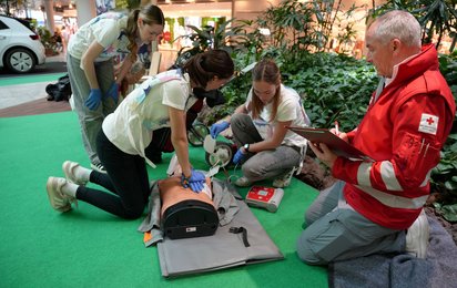 Eine Schülerin führt an einer Übungspuppe eine Herzdruck-Massage durch, während eine weitere Schülerin die Kontakte für den Defibrillator vorbereitet. Eine dritte Schülerin beugt sich über ihre Kolleginnen. Im Vordergrund beobachten ein Rotkreuz-Sanitäter die Arbeit der Schülerinnen.