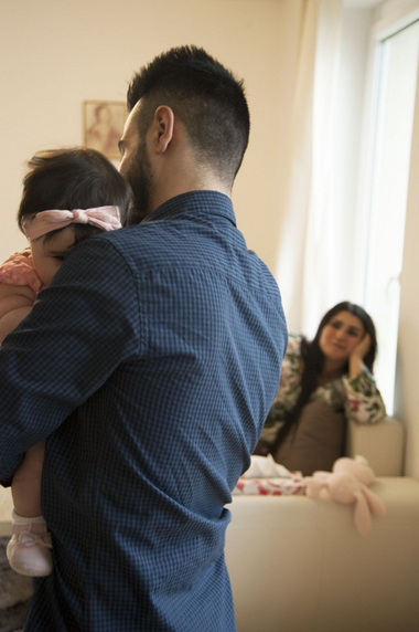 ein Vater ist von hinten zu sehen und hält das Kleinkind am Arm. Im Hintergrund sieht man eine erschöpfte Mutter