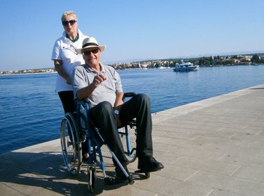 Ein Mann mit hellem Hut sitzt im Rollstuhl und zeigt lachend Richtung Kamera. Hinter ihm steht eine Rotkreuz-Mitarbeiterin und hält den Rollstuhl. Im Hintergrund sieht man das Meer und einen Hafen.