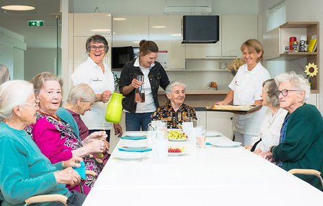 Eine Gruppe älterer Menschen sitzt gemeinsam lachend am Tisch und wird von drei Rotkreuz-Mitarbeiterinnen umsorgt.