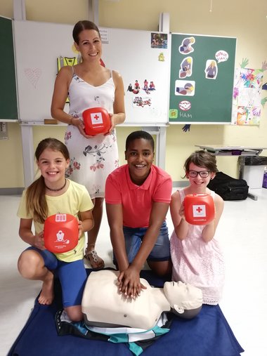 Ein Gruppenfoto von zwei Schülerinnen und einem Schüler mit ihrer Lehrerin. Man sieht eine Puppe, an der die Herzdruckmassage geübt werden kann.