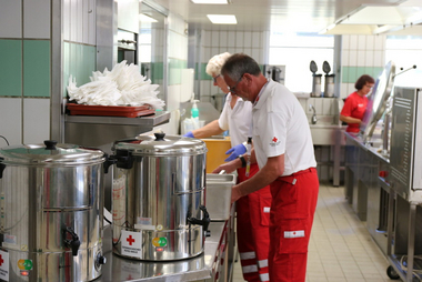 Zwei Rotkreuzmitarbeiter bereiten in einer Großküche Essen zu