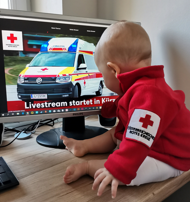 Säugling mit Uniformjacke sitzt vor dem Monitor auf dem der Livestream der Bezirksversammlung zu sehen ist