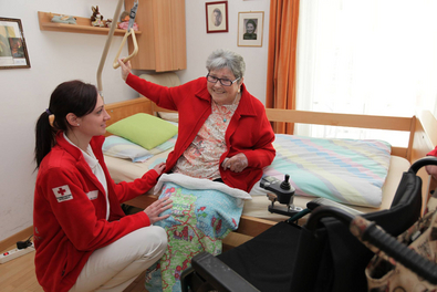 Eine ältere Dame sitzt aufrecht auf ihrem Pflegebett, eine Rotkreuz-Mitarbeiterin hockt vor ihr und unterstützt sie beim Aufstehen.