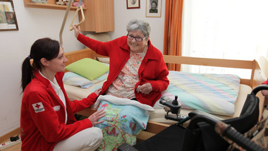 Eine ältere Dame sitzt aufrecht auf ihrem Pflegebett, eine Rotkreuz-Mitarbeiterin hockt vor ihr und unterstützt sie beim Aufstehen.