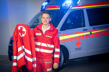 Rotkreuz-Mitarbeiter in Uniform vor einem Rettungswagen hat seine passende Jacke in der Hand.