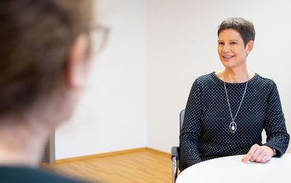 Eine Frau in Zivil sitzt am Tisch und spricht mit einer anderen Person, die man allerdings nur von hinten sieht