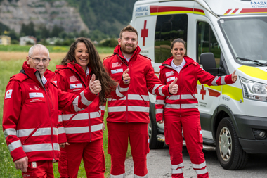 Vier Mitarbeiter des Roten Kreuzes Tirol in Roter Uniform stehen am Feld mit dem Rettungsauto im Hintergrund und heben ihren Daumen in die Höhe