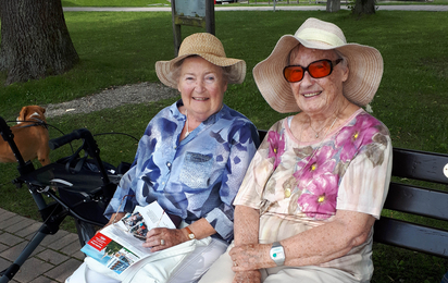 Zwei ältere Damen mit Sonnenhut sitzen lachend auf einer Sitzbank in einem Park.