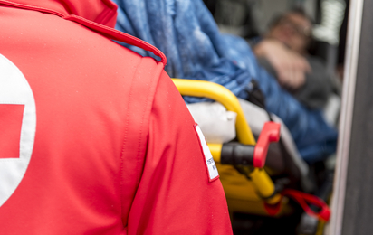 Zu sehen ist der Rücken eines Sanitäters, der einen Patienten auf der Trage ins Rettungsauto schiebt