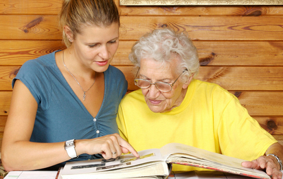 Junge Frau besucht ältere Dame, sie sitzen an einem Tisch und sehen sich gemeinsam ein Fotoalbum an. Die Seniorin trägt eine Brille.