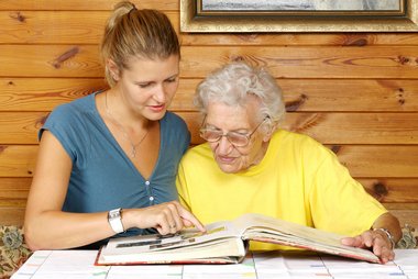 Junge Frau besucht ältere Dame, sie sitzen an einem Tisch und sehen sich gemeinsam ein Fotoalbum an. Die Seniorin trägt eine Brille.