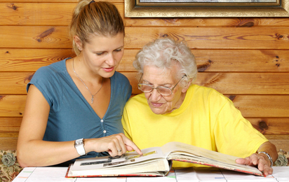 Junge Frau besucht ältere Dame, sie sitzen an einem Tisch und sehen sich gemeinsam ein Fotoalbum an. Die Seniorin trägt eine Brille.