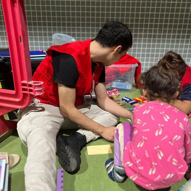 Ein Rotkreuz-Mitarbeiter spielt mit kleinen Kindern in einem Notquartier. Sie sitzen auf dem Boden.