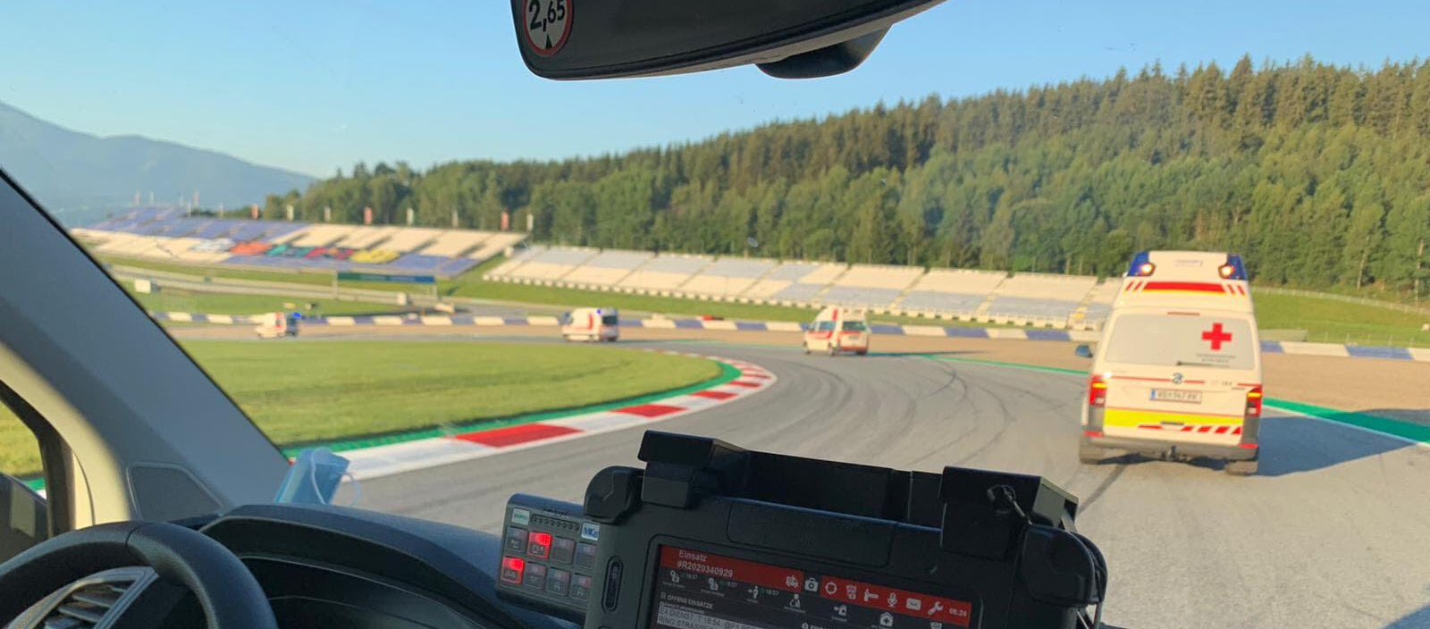 Einige Rettungswagen befinden sich auf einer Rennstrecke (dem Red Bull Ring) und beziehen grade ihre zugewiesenen Posten. Man sieht eine Fahrerkabine von Innen und weitere Rettungswagen vor dem Fahrzeug.