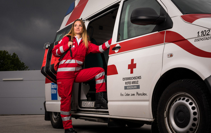 Eine junge Sanitäterin steht in der Tür eines Rettungsautos. Am Rücken trägt sie einen Notfallrucksack.