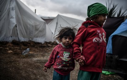 © ICRC / Abed Zaqout Zwei kleine Kinder stehen Hand in Hand auf schlammigen Boden vor Zelten in Gaza.