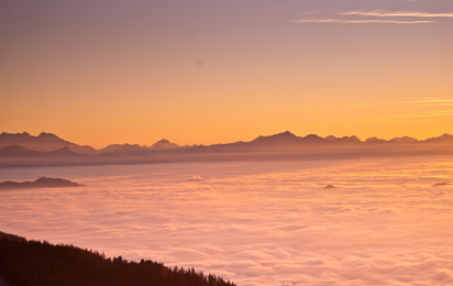 Man sieht einen wunderschönen Sonnenuntergang über einer Wolkendecke und Bergen im Hintergrund.