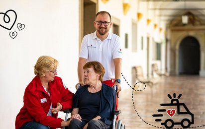 Eine ältere Frau sitzt im Rollstuhl und wird von zwei Rotkreuz-Mitarbeiter:innen betreut.