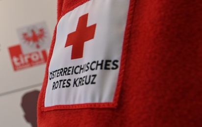 Zu sehen ist ein Teil einer roten Rotkreuz Fleecejacke mit dem Rotkreuz Logo. Im Hintergrund sieht man verschwommen das Logo des Landes Tirol