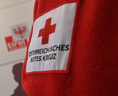 Zu sehen ist ein Teil einer roten Rotkreuz Fleecejacke mit dem Rotkreuz Logo. Im Hintergrund sieht man verschwommen das Logo des Landes Tirol