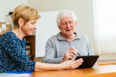 Ein älterer Herr mit fröhlichen Gesicht, welcher gemeinsam mit einer Dame ein Tablet bedient.