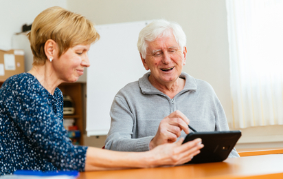 Ein älterer Herr mit fröhlichen Gesicht, welcher gemeinsam mit einer Dame ein Tablet bedient.