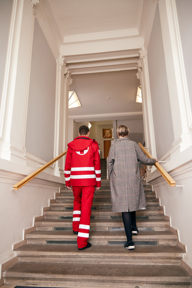 Männlicher Rotkreuz-Mitarbeiter in Uniform und eine weibliche Person gehen die Stufen eines Wohnhauses hoch.