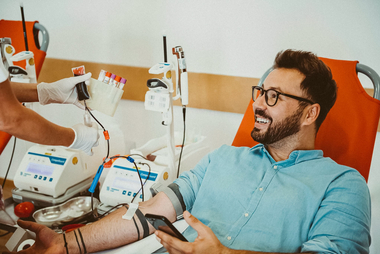 Junger Mann beim Blutspenden mit Brille, Tattoos und Handy