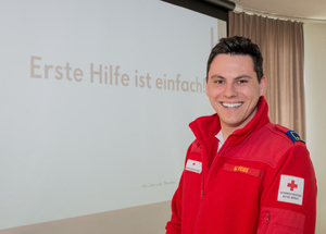 Weiss Moritz - Erste-Hilfe-Lehrbeauftragter - steht in einem Lehrsaal. Im Hintergrund sieht man den Spruch "Erste Hilfe ist einfach".