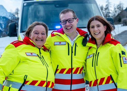 Drei Sanitäterinnen stehen nebeneinander mit einer gelben Uniform Oberteil. Sie stehen vor einem Auto und im Freien