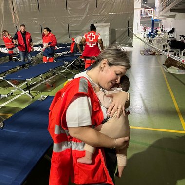 Eine Rotkreuz-Mitarbeitern hält in einem Notquartier im Baby im Arm. Im Hintergrund sind Feldbetten zu sehen.