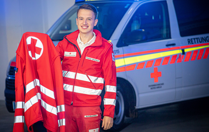 Rotkreuz-Mitarbeiter in Uniform vor einem Rettungswagen hat seine passende Jacke in der Hand.