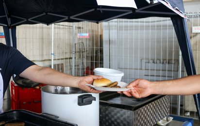 Ein Teller mit einer Semmel und mehreren Bratwürsten wird von einer Person an eine andere übergeben.