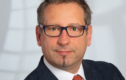 Portrait eines Mannes mit kurzen Haare, dunkler Brille und dunkelgrauem Sakko.