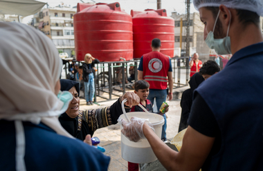 Eine syrische Frau bekommt einen Kübel Wasser von einem Mitarbeiter des Syrisch-Arabischen Roten Halbmonds. Im Hintergrund sieht man Wassertanks.