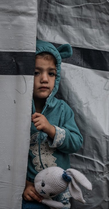 Ein kleines Mädchen in Gaza schaut vorsichtig hinter einem Zelt hervor und hält ihren schmutzigen Stoffhasen in der Hand.