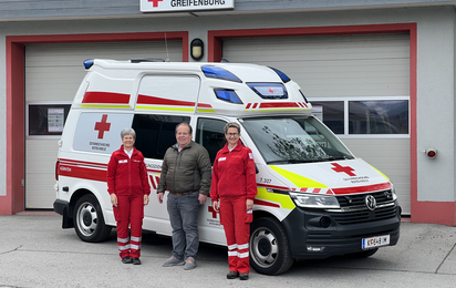 Gruppenfoto von drei Personen in Rotkreuz-Uniform vor einem Rettungswagen und einem Rotkreuz-Gebäude