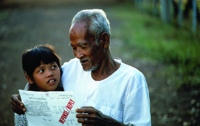 Ein älterer asiatischer Mann und ein junges Mädchen lesen gemeinsam Rotkreuz-Nachrichten. Der Mann umarmt das Kind dabei.