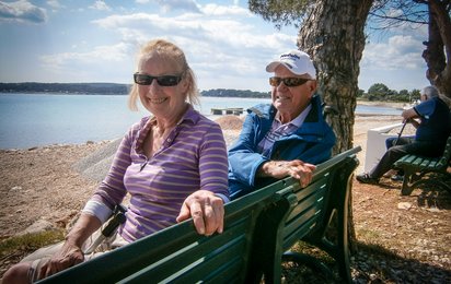 Ein älteres Paar sitzt auf einer Parkbank am Meer. Beide strahlen in die Kamera.