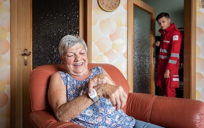 Eine alte Dame sitzt in ihrem Wohnzimmer und drückt den Kopf ihres Rufhilfe-Armbands und ein Rotkreuz-Sanitäter kommt von hinten durch die Tür um zu helfen.