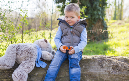 Ein kleiner Junge sitzt mit seinem Stoff-Teddybären auf einem Baumstamm.