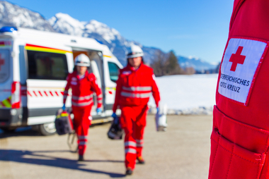 Zwei Sanitäter mit Gerätschaften gehen vom RTW Richtung eines weiteren Sanitäters, dessen Rotes Kreuz zu sehen ist.