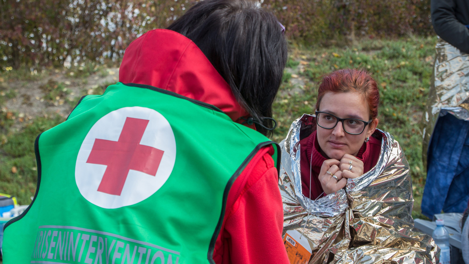 Eine Mitarbeiterin der Krisenintervention spricht mit einer Frau, die in einer Rettungsdecke eingewickelt ist. Sie bekommt psychosoziale Unterstützung während einer Krise