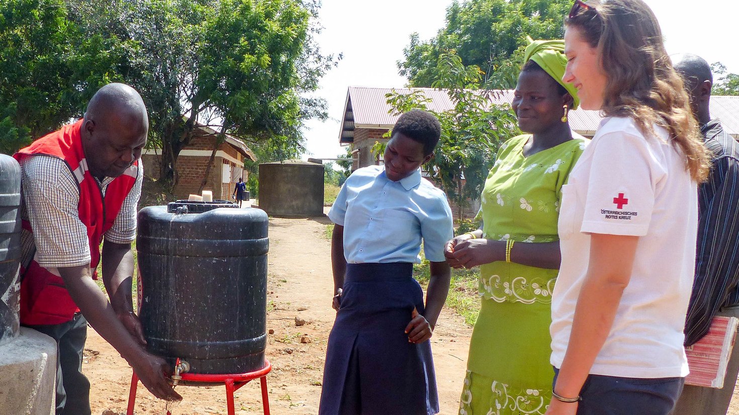 Eine Rotkreuz-Helferin in Uganda unterstützt bei einem Wasserprojekt. Ein lokaler Rotkreuz-Helfer wäscht sich die Hände unter einem Kanister, weitere Personen schauen zu.