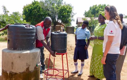 Eine Rotkreuz-Helferin in Uganda unterstützt bei einem Wasserprojekt. Ein lokaler Rotkreuz-Helfer wäscht sich die Hände unter einem Kanister, weitere Personen schauen zu.