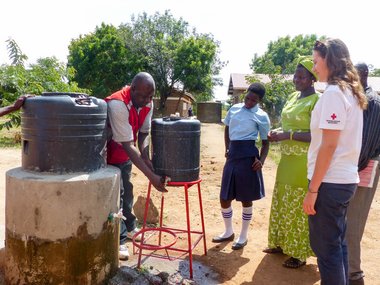 Eine Rotkreuz-Helferin in Uganda unterstützt bei einem Wasserprojekt. Ein lokaler Rotkreuz-Helfer wäscht sich die Hände unter einem Kanister, weitere Personen schauen zu.