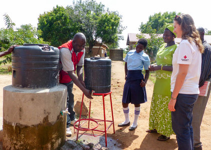 Eine Rotkreuz-Helferin in Uganda unterstützt bei einem Wasserprojekt. Ein lokaler Rotkreuz-Helfer wäscht sich die Hände unter einem Kanister, weitere Personen schauen zu.