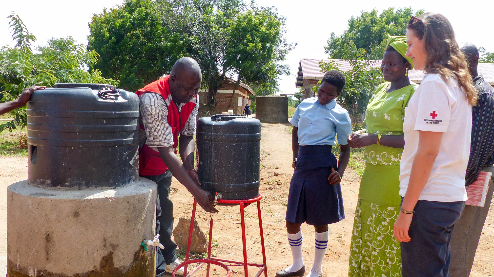 Eine Rotkreuz-Helferin in Uganda unterstützt bei einem Wasserprojekt. Ein lokaler Rotkreuz-Helfer wäscht sich die Hände unter einem Kanister, weitere Personen schauen zu.