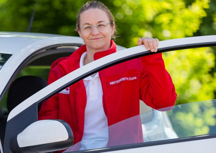 Eine Diplomierte Krankenpflegerin des Roten Kreuzes Burgenland steht in der geöffneten Autotür ihres Dienstautos und lächelt